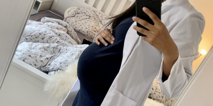 Frau, deren Kopf nicht zu sehen ist, mit weißem Kittel, schwarzem Kleid hält rechte Hand auf Babybauch, in der linken Hand hält sie ein Handy, im Hintergrund ein weißes Bett mit bunter Bettwäsche