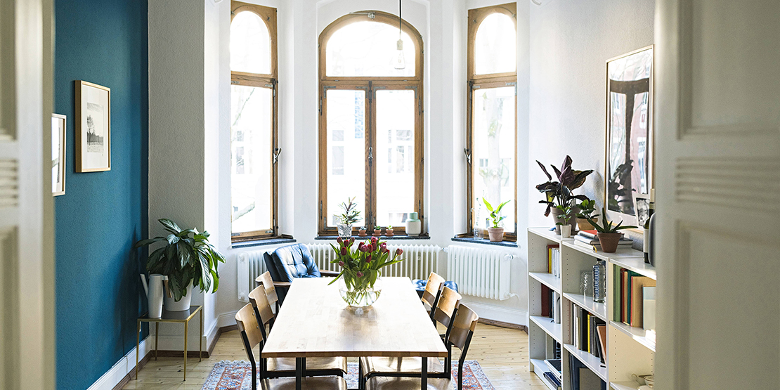 Wohnzimmer mit hohen Wänden, in der Mitte einer heller Holztisch mit sechs Stühlen, links eine türkisfarbene Wand, rechts eine weiße Wand davor eine niedriges Bücherregal,  im Hintergrund drei hohe Fenster in einem Erker.