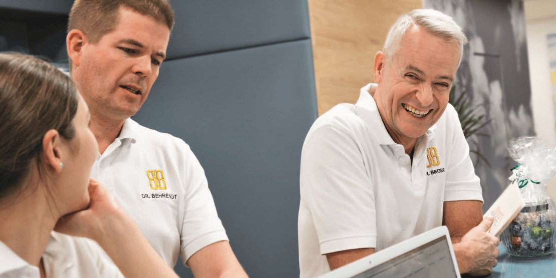 Zwei Ärzte und eine Ärztin stehen entspannt an einem Empfangstresen einer Arztpraxis und lachen herzlich.