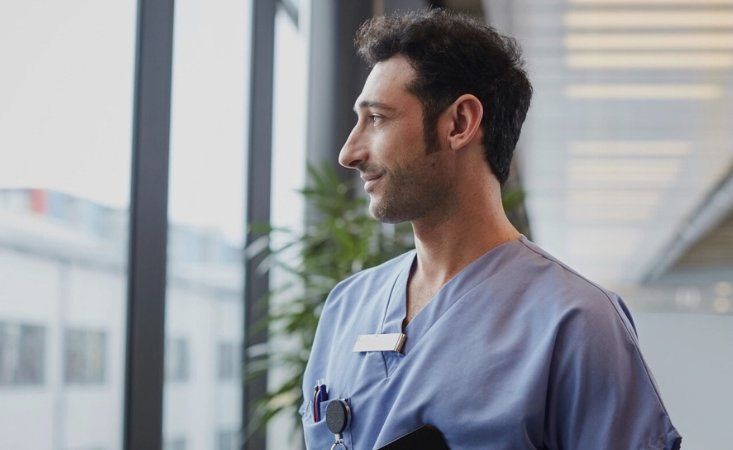 Dunkelhaariger Mann mit blauem Kasack schaut aus dem Fenster.