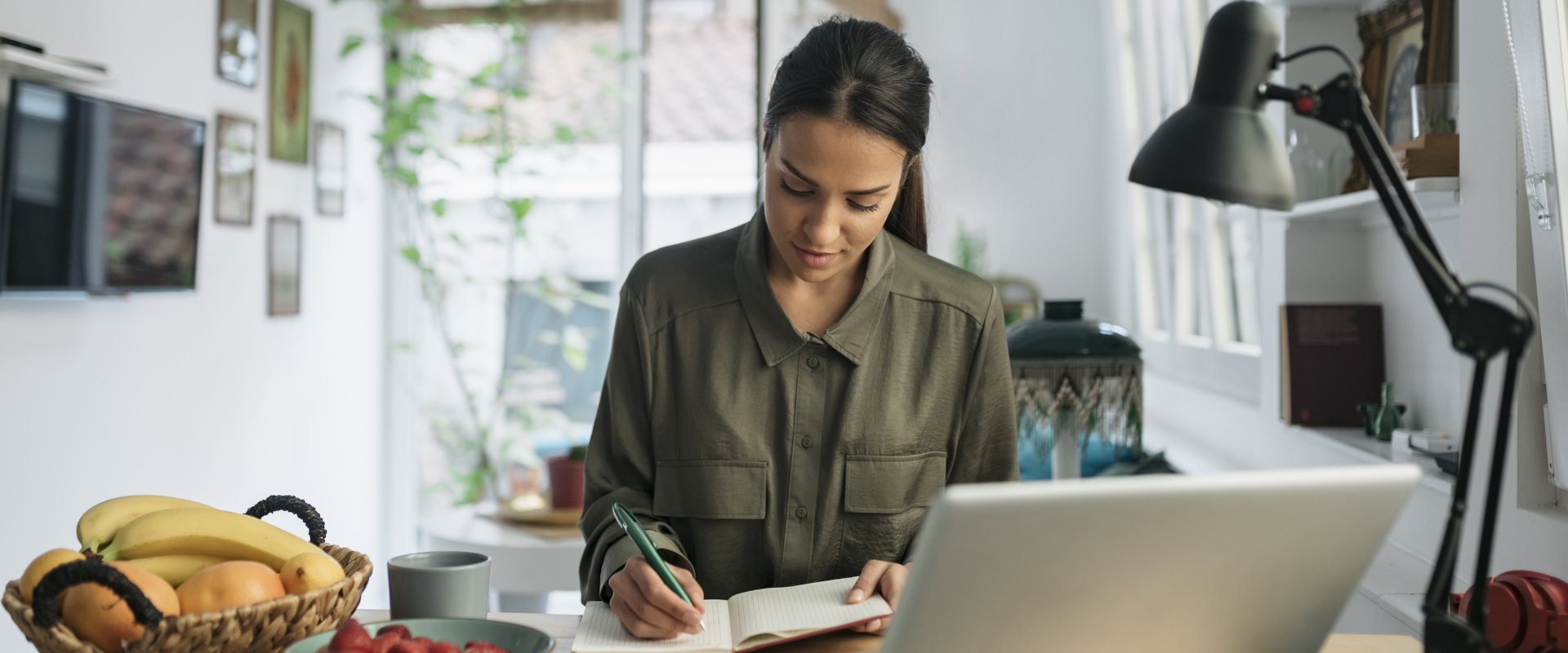 Junge dunkelhaarige Frau, die vor einem Laptop sitzt, schreibt in ein Heft