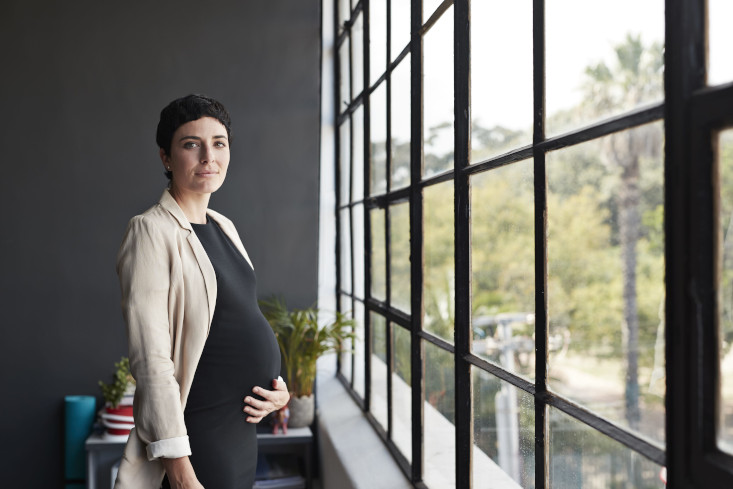vor einem modernen Glasfenster mit schwarzen Sprossen steht ein Frau mit schwarzem Kleid und beiger Jacke, die mit ihrer linken  Hand de Babybauch hält und in die Kamera schaut