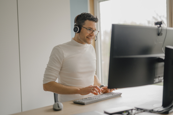 Ein junger Mann mit Brille steht an seinem Schreibtisch und führt ein Online-Meeting.
