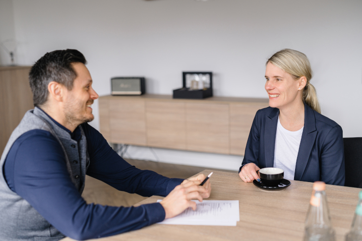 Ein Mann und eine Frau mittleren Alters sitzen sich an einem Tisch gegenüber und unterhalten sich lächelnd. Die Frau sitzt vor einer Tasse und der Mann hat Unterlagen vor sich liegen.