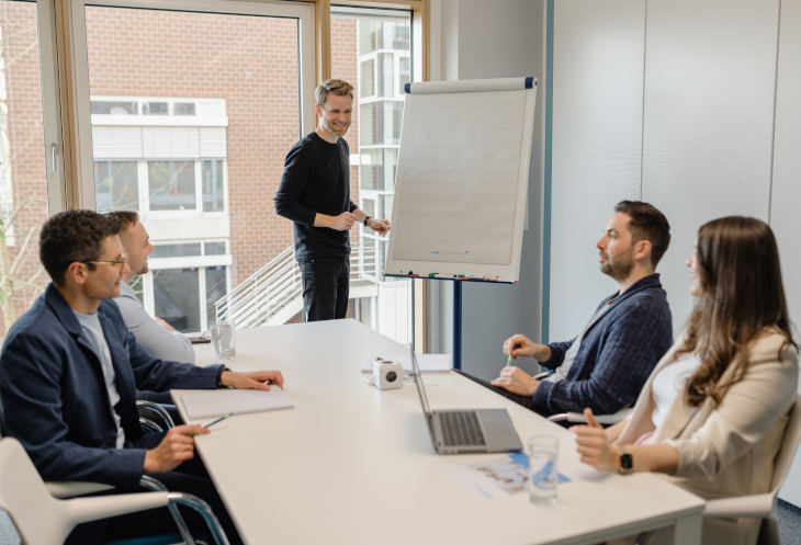 Vier Personen sitzen in einem Meetingraum an einem Tisch. Ein junger Mann steht an einem Flipchart.