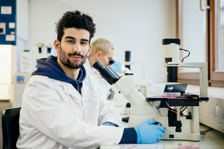 Junger dunkelhaariger Medizinstudent sitzt vor einem Mikroskop und schaut in die Kamera. Er trägt über seinem Hoodie einen weißen Kittel.