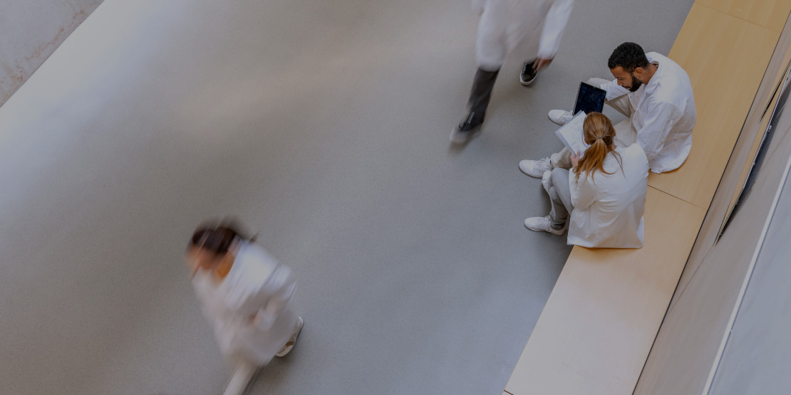 Verschwommenes Bild von Menschen in Bewegung und mit Kitteln, zwei, eine Frau und ein Mann  sitzen auf beiger Holzbank