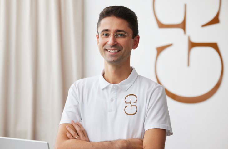 Ein Mann mit weißem Polohemd steht mit verschränkten Armen dar und lächelt in die Kamera