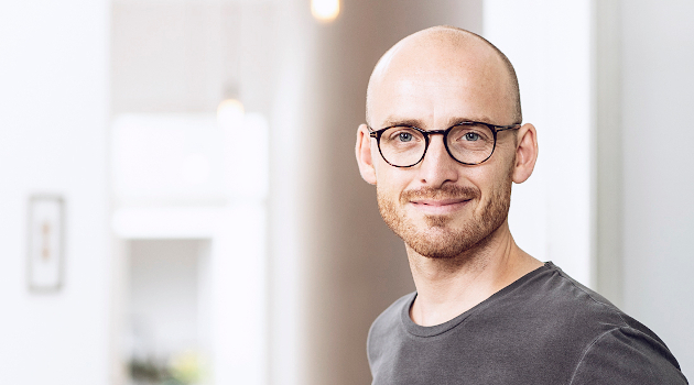 Ein Mann Mitte 30 schaut die Betrachter freundlich an. Er trägt eine Brille und ein graues T-Shirt.