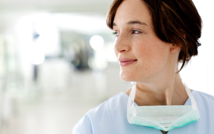 Eine junge Ärztin lehnt lächelnd an der Wand im Flur eines Krankenhauses. Sie trägt einen Kittel und um ihren Hals hängt eine OP-Maske.