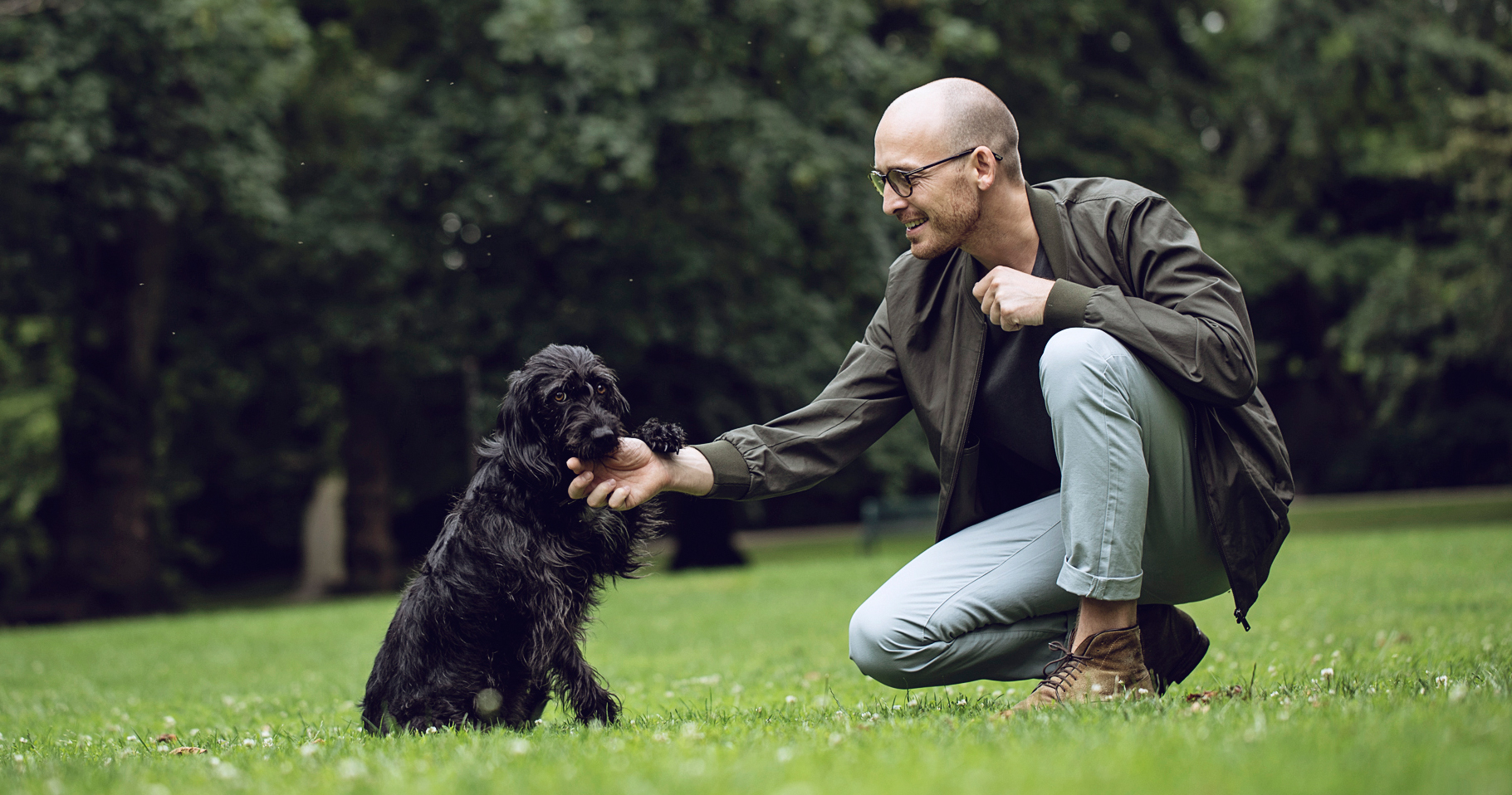 Ein junger Mann kniet auf einer Wiese im Park und streichelt lächelnd einen mittelgroßen, schwarzen Hund. 