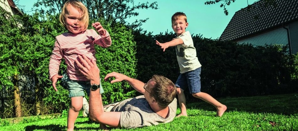 Ein Vater liegt im Gras. Er spielt mit seinen beiden kleinen Kindern, einem Jungen und einem Mädchen.